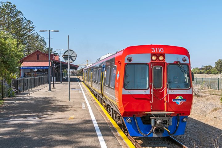 A train passing through the Outer Harbor Station. It is red and blue with the Adelaide Metro logo on the bottom right.