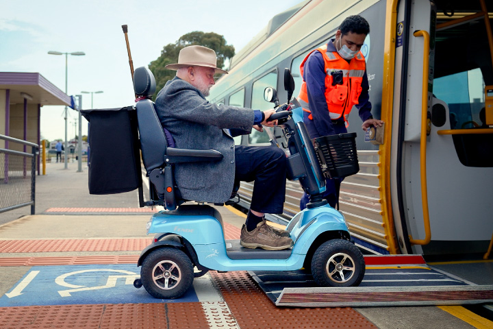 A passenger in a mobility scooter boarding a train. A public transport worker is assisting the passenger onto the boarding ramp.